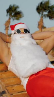 Noel Baba yaz tatili. Noel Baba güneşleniyor. Komik Noel Baba, güneş gözlükleriyle ve paletleriyle oturuyor, dinleniyor, deniz kenarında, palmiye ağaçlarına yaslanıyor..