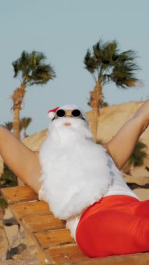 Noel Baba yaz tatili. Noel Baba güneşleniyor. Komik Noel Baba, güneş gözlükleriyle ve paletleriyle oturuyor, dinleniyor, deniz kenarında, palmiye ağaçlarına yaslanıyor..