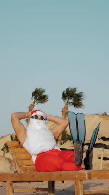 Noel Baba yaz tatili. dikey video. Noel Baba güneşleniyor. Komik Noel Baba, güneş gözlükleriyle ve paletleriyle oturuyor, dinleniyor, deniz kenarında, palmiye ağaçlarına yaslanıyor..