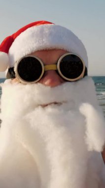 Noel Baba blogcusu. Noel Baba selfie çeker. Güneş gözlüklü komik Noel Baba, sahilde, palmiye ağaçlarının önünde selfie videosu çekiyor. Noel Baba yaz tatili.