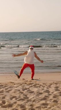 Noel Baba yaz tatili. Noel Baba eğleniyor. Komik Noel Baba, güneş gözlüğü ve yüzgeçleriyle deniz kıyısındaki kumlu sahilde dans edip zıplıyor. Yüzmek için acele ediyor, şnorkel.