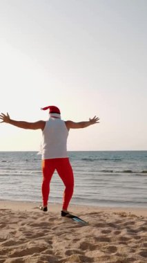 Noel Baba yaz tatili. Noel Baba eğleniyor. Komik Noel Baba, paletlerle kumlu sahilden denize koşuyor. Yüzmek için acele ediyor, şnorkel. Arkadan bak. dikey video