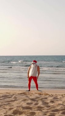 Noel Baba yaz tatili. Noel Baba eğleniyor. Komik Noel Baba, güneş gözlüğü ve yüzgeçleriyle deniz kıyısındaki kumlu sahilde dans ediyor. dikey video