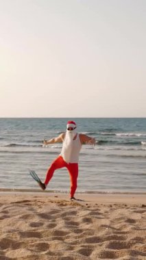 Noel Baba yaz tatili. Noel Baba eğleniyor. Komik Noel Baba, güneş gözlüğü ve yüzgeçleriyle deniz kıyısındaki kumlu sahilde dans edip zıplıyor. Yüzmek için acele ediyor, şnorkel. dikey video