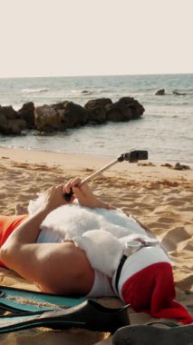 Noel Baba blogcusu. Noel Baba selfie çeker. Güneş gözlüklü komik Noel Baba, deniz kenarındaki kumlu sahilde uzanırken selfie videosu çekiyor. Noel Baba yaz tatili.