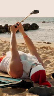Noel Baba blogcusu. Noel Baba selfie çeker. Güneş gözlüklü komik Noel Baba, deniz kenarındaki kumlu sahilde uzanırken selfie videosu çekiyor. Noel Baba yaz tatili.