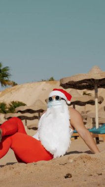 Noel Baba yaz tatili. Noel Baba eğleniyor. Koruyucu maskeli, güneş gözlüklü ve paletli komik Noel Baba deniz kenarında palmiye ağaçlarının arasında uzanırken dinleniyor.