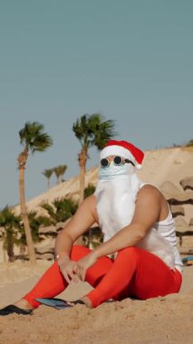Noel Baba yaz tatili. Noel Baba eğleniyor. Koruyucu maskeli, güneş gözlüklü ve paletli komik Noel Baba deniz kenarında palmiye ağaçlarının arasında uzanırken dinleniyor.