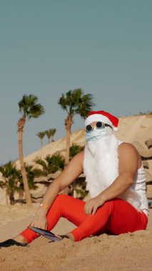 Noel Baba yaz tatili. Noel Baba eğleniyor. Koruyucu maskeli, güneş gözlüklü ve paletli komik Noel Baba deniz kenarında palmiye ağaçlarının arasında uzanırken dinleniyor.