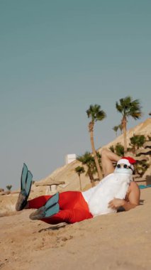 Noel Baba yaz tatili. Noel Baba eğleniyor. Koruyucu maskeli, güneş gözlüklü ve paletli komik Noel Baba deniz kenarında palmiye ağaçlarının arasında uzanırken dinleniyor.