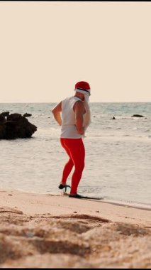 Noel Baba yaz tatili. Noel Baba eğleniyor. Komik Noel Baba, güneş gözlüğü ve yüzgeçleriyle kumlu sahilden yavaşça deniz suyuna giriyor ve yüzmek üzere..