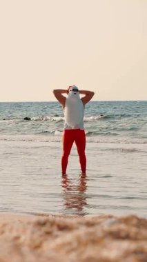 Noel Baba güneşleniyor. Noel Baba eğleniyor. Güneş gözlüklü komik Noel Baba, sahildeki deniz suyunda duruyor. Noel Baba yaz tatili