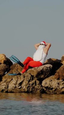Noel Baba güneşleniyor. Noel Baba eğleniyor. Komik Noel Baba, güneş gözlüğü ve yüzgeçleriyle deniz kıyısındaki kayalık sahilde oturuyor. Noel Baba yaz tatili