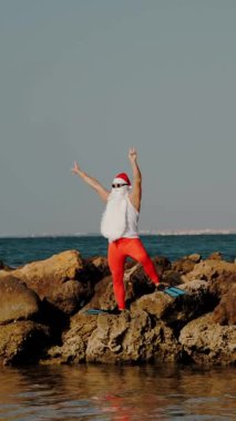 Noel Baba yaz tatili. Noel Baba eğleniyor. Komik Noel Baba, güneş gözlüğü ve yüzgeçleriyle deniz kıyısındaki kayalık sahilde dans ediyor. dikey video.