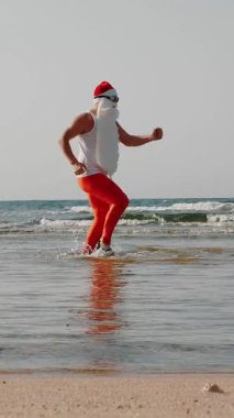 Komik Noel Baba. Noel Baba, güneş gözlüğü ve paletlerle kumsalın yanındaki deniz suyunda eğleniyor. Noel Baba yaz tatili.