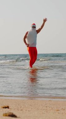 Komik Noel Baba. Noel Baba, güneş gözlüğü ve yüzgeçleriyle el sallayarak kumsalın yanındaki deniz suyunda eğleniyor. Noel Baba yaz tatili.