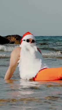 Komik Noel Baba. Noel Baba, güneş gözlüğü ve paletlerle oturup eğleniyor kumsalın yanındaki deniz suyunda dinleniyor. Noel Baba yaz tatili