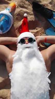 Güneş gözlüğü takan Noel Baba deniz kenarındaki kumlu sahilde güneşleniyor. Üst Manzara. Noel Baba yaz tatili.