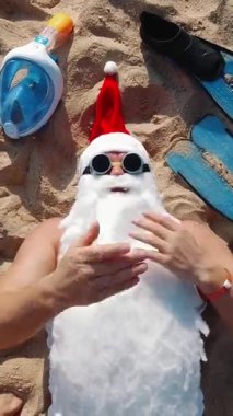 Güneş gözlüğü takan Noel Baba deniz kenarındaki kumlu sahilde uzanıyor ve selfie video kamerasına öpücük yolluyor. Üst Manzara. Noel Baba yaz tatili. Noel Baba blogcusu.