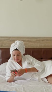 - Otel odasında. Kafasına havlu bağlanmış beyaz bornozlu genç bir kadın, bir otel odasında büyük bir yatakta dinlenirken, akıllı telefon kullanıyor.