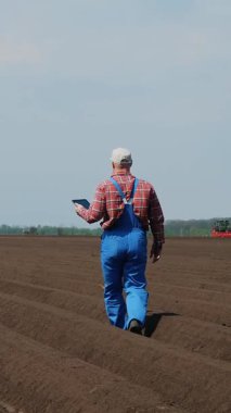Çiftçi, tarım uzmanı tarlada özel toprak sıralarıyla yürüyor. Test ediyor, tablet kullanıyor, ekicinin patates ekme kalitesini ölçüyor. Modern tarımsal yetiştirme. Bahar güneşli gün.