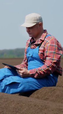 Tarımcı çiftçi, tarlada özel toprak sıralarının arasında oturur. Test ediyor, tablet kullanıyor, ekicinin patates ekme kalitesini ölçüyor. Modern tarımsal olarak patates yetiştirme. Bahar güneşli gün.