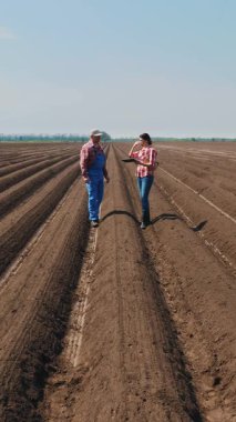 Çiftçi ve tarımcı, tarladaki toprak sıraları arasında tabletle, ekicinin patates ekme kalitesini test ederek derin bir çizgide yürüyor. Modern tarım. Bahar güneşli gün.