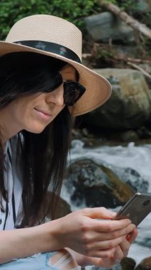 Ormanda nehirde yürüyüş yapan akıllı telefondan bakan kıvırcık bir kızın portresi. Dağ yürüyüşünde genç bir kadın Summer 'da haritada rota bulmak için telefondaydı. Turizm, seyahat, yürüyüş, gezi, macera konsepti.