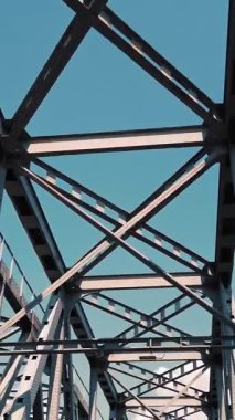 Metal köprü. Mavi gökyüzüne karşı kaba demir yapılar, hareket halindeki bir arabadan çekiliyor. Alt manzara. Nehrin karşısındaki demiryolu ve yol köprüsü. Çelik kirişleri aşan siluet.