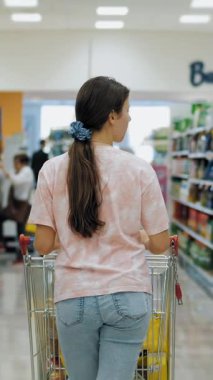 Süpermarkette alışveriş arabası. Kız süpermarkette koridorlar arasında alışveriş arabasını yuvarlıyor. Arkadan bak. Genç kız seçiyor, alacak bir şeyler arıyor, süpermarkette yürüyor.