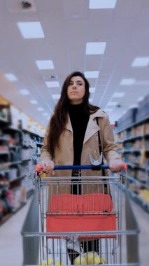 Çeşitli ürünlerle dolu raflarla çevrili bir süpermarket koridorunda alışveriş arabasını iten genç bir kadının görüntüsü. Odaklanmış görünüyor ve modaya uygun bir kıyafet giyiyor.