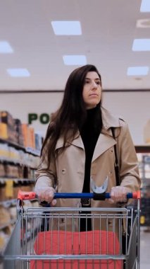 Genç bir kadın, kırmızı bir alışveriş arabasını iterek bir süpermarket koridorunda rahatça geziniyor. Şık bej bir trençkot giyiyor, market seçeneklerini araştırıyor.