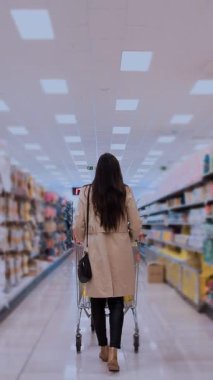 Perakende mağazasında ev eşyalarıyla dolu bir koridorda yürürken bir kadın arkadan görülüyor. Sıradan giyinmiş ve bir çanta taşıyor. Temiz ve düzenli bir ortamda alışveriş yapıyor.