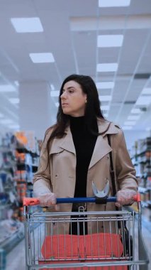 Trençkot giymiş bir kadın, bir alışveriş arabasını süpermarket koridoruna itiyor, çeşitli ürünleri dikkatlice düşünüyor. Odaklanmış ifadesi, perakende ortamında tüketici davranışlarını ve karar verme sürecini yansıtıyor.