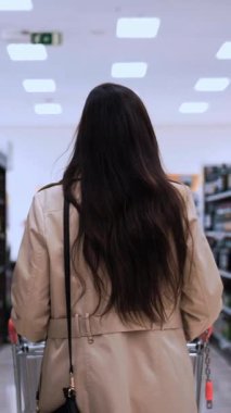 Uzun siyah saçlı, bej renkli trençkotlu bir kadın bir alışveriş arabasını süpermarket şarap reyonuna itiyor.