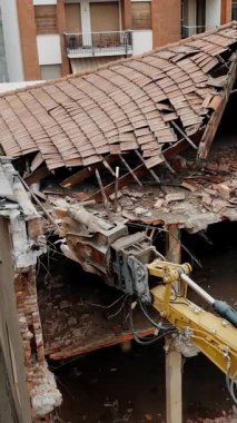 The photo shows a heavy-duty excavator actively demolishing a severely damaged and dilapidated building in an urban setting, emphasizing the ongoing urban renewal efforts