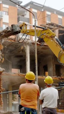 Güvenlik kaskı ve yansıtıcı yelek giyen iki inşaat işçisi, çok katlı bir binanın yıkımına tanık oluyor. Hidrolik öğütücüyle donatılmış büyük sarı bir kazıcı.