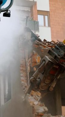 Güçlü bir hidrolik kazıcı binanın bir kısmını enkaz ve tozla yıkıyor ve tipik bir kentsel yenilenme sahnesi sergiliyor.
