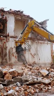 Güçlü bir kazıcı, eski bir binayı enkaz gibi yıkıyor. Güvenlik ekipmanları kullanan bir işçi tarafından yakından denetleniyor ve kentsel gelişimi gösteriyor.