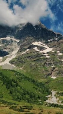 Yeşil vadilerle ve açık mavi gökyüzünün altında akan bir akarsula karışmış bulutlu zirvelerle nefes kesici bir sıradağ manzarası.