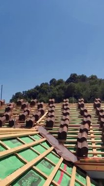 Tepenin üzerine yeşil brandası olan bir çatı inşa ediliyor. Şantiye, ağaçlar ve tepelerle çevrili kırsal bir alanda yer alıyor.