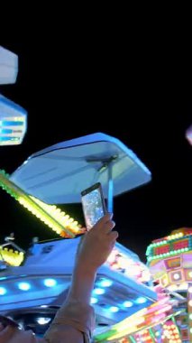 Kadın eli akıllı telefon tutuyor ve fotoğraf çekiyor ya da video çekiyor. Işıklı atlıkarınca. Antika atlıkarıncanın inanılmaz renkli yanıp sönen ışığı. Popüler Sandalye Eğlence Parkı 'nda Sallanıyor