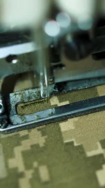 Askeri kıyafetler kamufle edildi. Dikiş. Yakın plan. Terzi dikiş atölyesinde dikiş makinesi kullanarak kamuflaj kumaşına düğme deliği dikiyor. Haki, koyu yeşil kumaş..
