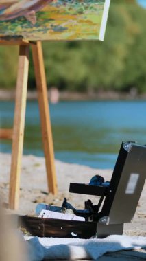 Yakın çekim, nehrin kıyısında kumlu bir sahil, kumsalda bir resim sehpası duruyor, ve yanında farklı renklerde tüpler olan özel bir bavul var..