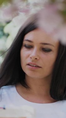 Beyaz elbiseli güzel bir kadın ressamın portresi, ressam, çiçek açan bahar elma bahçesinde bir resim çizer, özel bir çizim tekniği kullanarak tuvale özel küçük bir spatula ile boya uygular. Boyalı bir palet tutar.