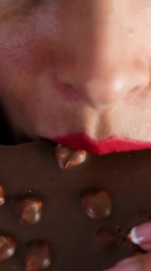 Tatlı cazibe. Kırmızı dudaklı bir kadının çikolatayı ısırması, fındıklı çikolata yiyen güzel esmer bir kadının dudakları.