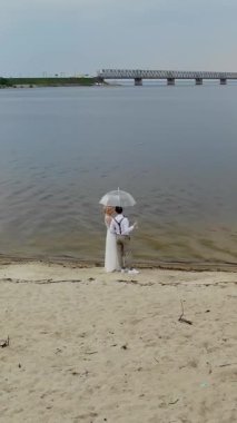 aero, güzel yeni evliler sahilde duran, şampanya bardak ve şeffaf bir şemsiye altında, mavi gökyüzü, nehir ve nehir üzerinde büyük bir köprü karşı. bahar günü. Düğün