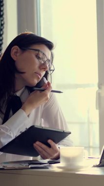 Odaklanmış, başarılı, tablet ve dizüstü bilgisayarı olan başarılı bir iş kadını ya da sekreteri olan genç bir kadın, ekrana bakıyor, ofisteki akıllı telefondan arıyor, müşteriye danışıyor. İş, teknoloji, iletişim ve insan kavramı