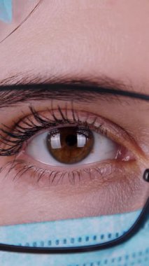 Yakın plan, göz, gözlüklü, mavi tıbbi maskeli ve tıbbi şapkalı kadın doktor yüzü..