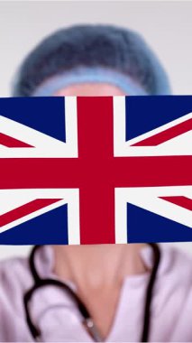 Yakın plan. Gözlüklü doktor, mavi tıbbi şapkalı, eldivenli, İngiltere bayrağıyla tıbbi maske takıyor. Doktorlar, koronavirüs sırasında devletin koruması, küresel salgın. Kavram